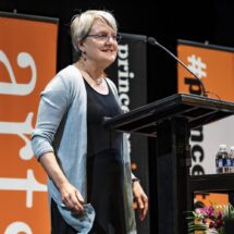 Stacy Wolf, seen at 3/4 view, stands smiling behind a podium on stage by black and orange University banners.