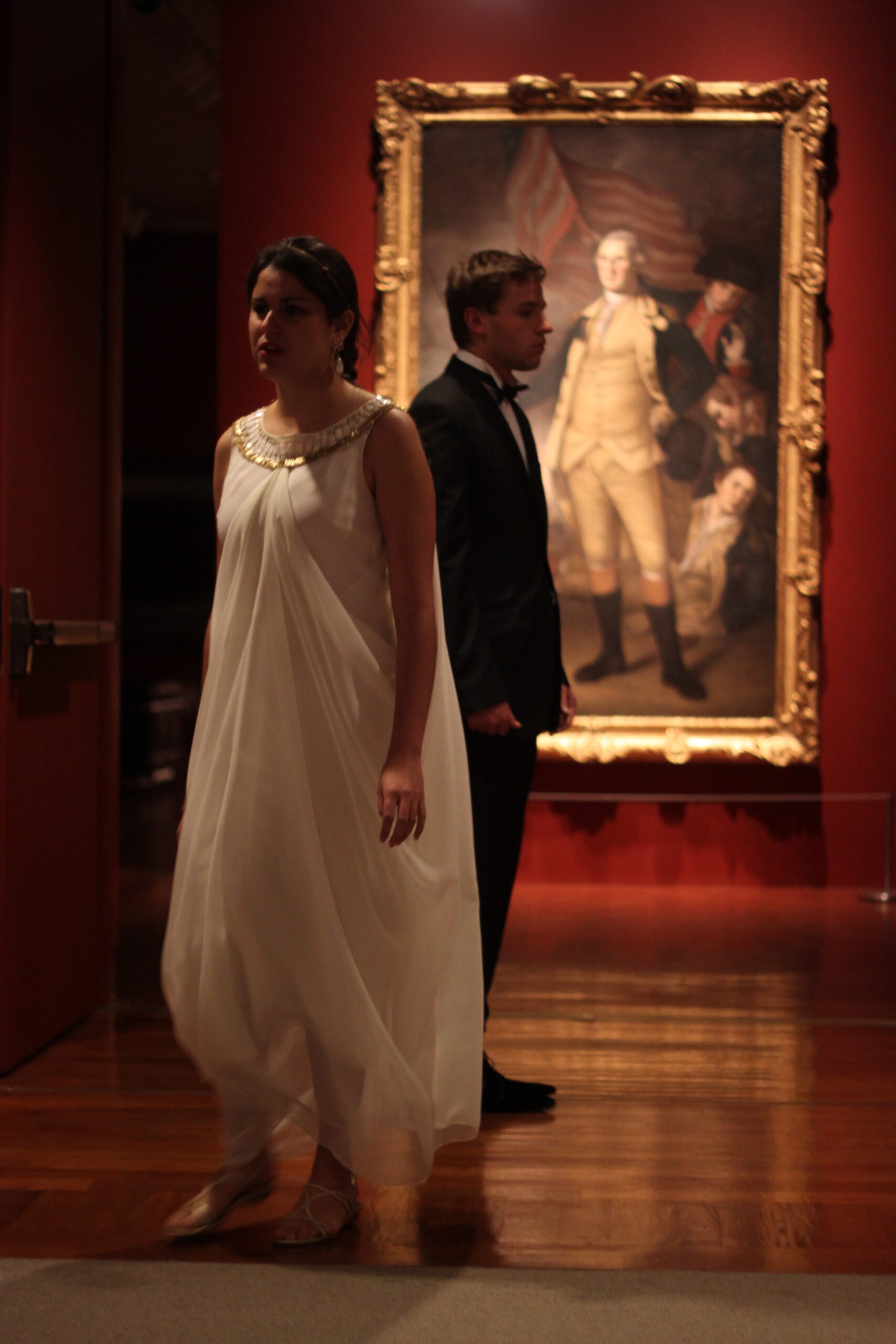 Two people dressed in formal wear stand apart looking opposite directions in a gallery space with a large painting and red walls.