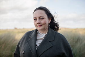 Caitríona stands outdoors under a cloudy grey-blue sky. She wears a grey wool coat and her hair is blown back by the wind.