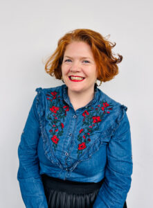 Jen Coppinger smiles widely. She has short red wavy hair and wears a denim shirt embroidered with red flowers.