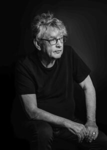 Paul Muldoon, dressed in black, sits leaning with his right arm on right knee while he looks off to his left.