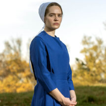 Person in a blue dress and white cap standing outdoors against autumn trees.