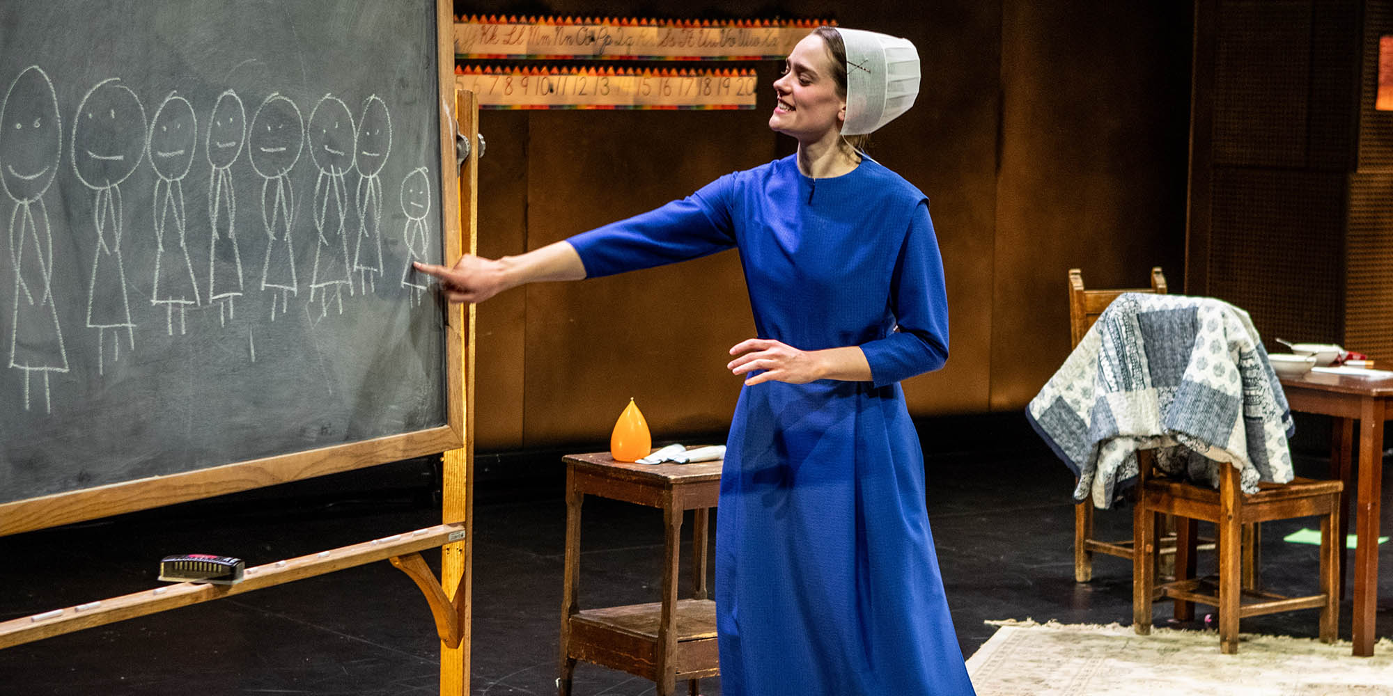 A woman in a blue dress points at stick figure drawings on a chalkboard.