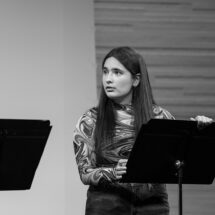 Kaelani stands behind a music stand, looking at another actor on her right.