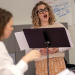 A woman singing with a music stand in a room with posters on the wall as someone conducts.