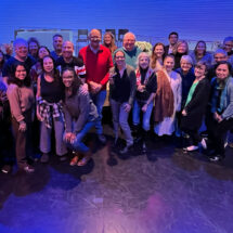 A large group of people crowd together for a portrait while standing in a blue-lit studio space