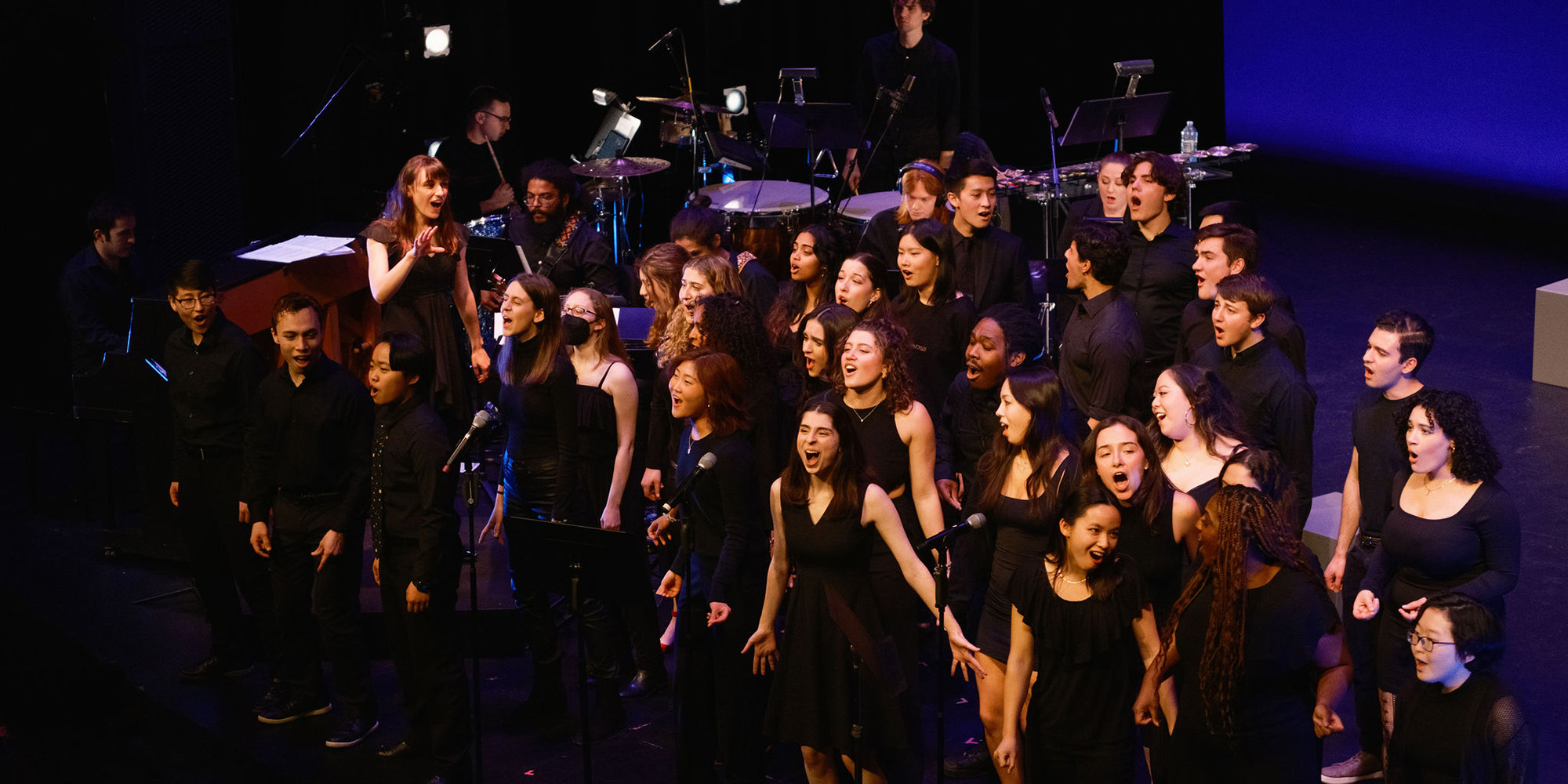 a chorale group wearing black stands scattered onstage, singing and smiling.