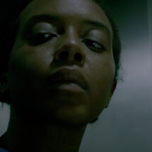 A woman's face seen close up in shadow. Her expression is guarded and possibly afraid.