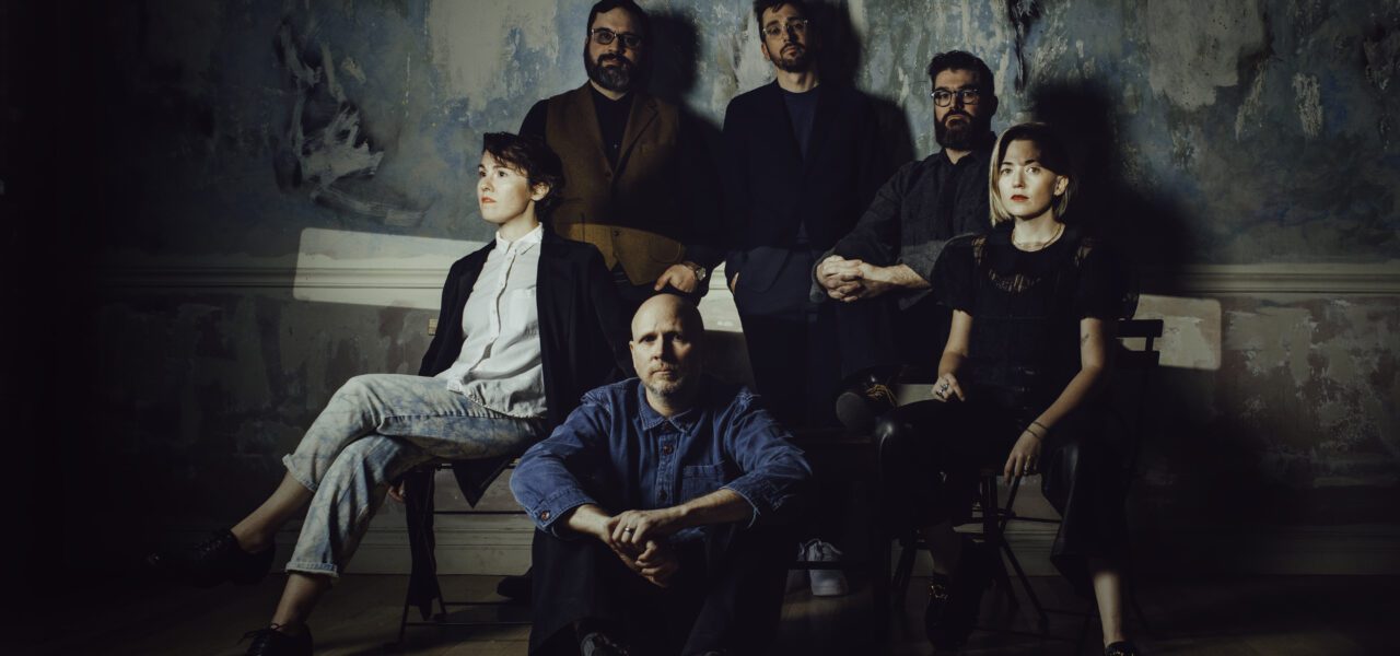 Musicians So Percussion and Caroline Shaw sit in a casual group photo in a shadowed interior room.