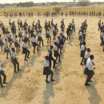 A large crowd of people dance in a series of lines out on a dusty plain landscape