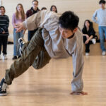 Person breakdancing in front of a group of onlookers in a classroom setting.