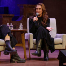 Two women sitting in chairs holding microphones have a discussion on stage.