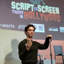 Man speaking on stage with a presentation behind him titled 