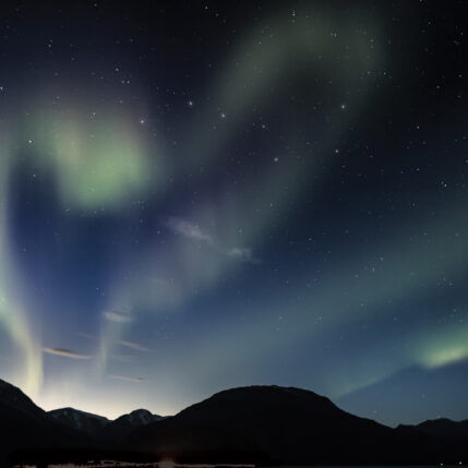 Green aurora borealis lights in a dark sky form the shape of a heart over a dark mountain landscape silhouette.