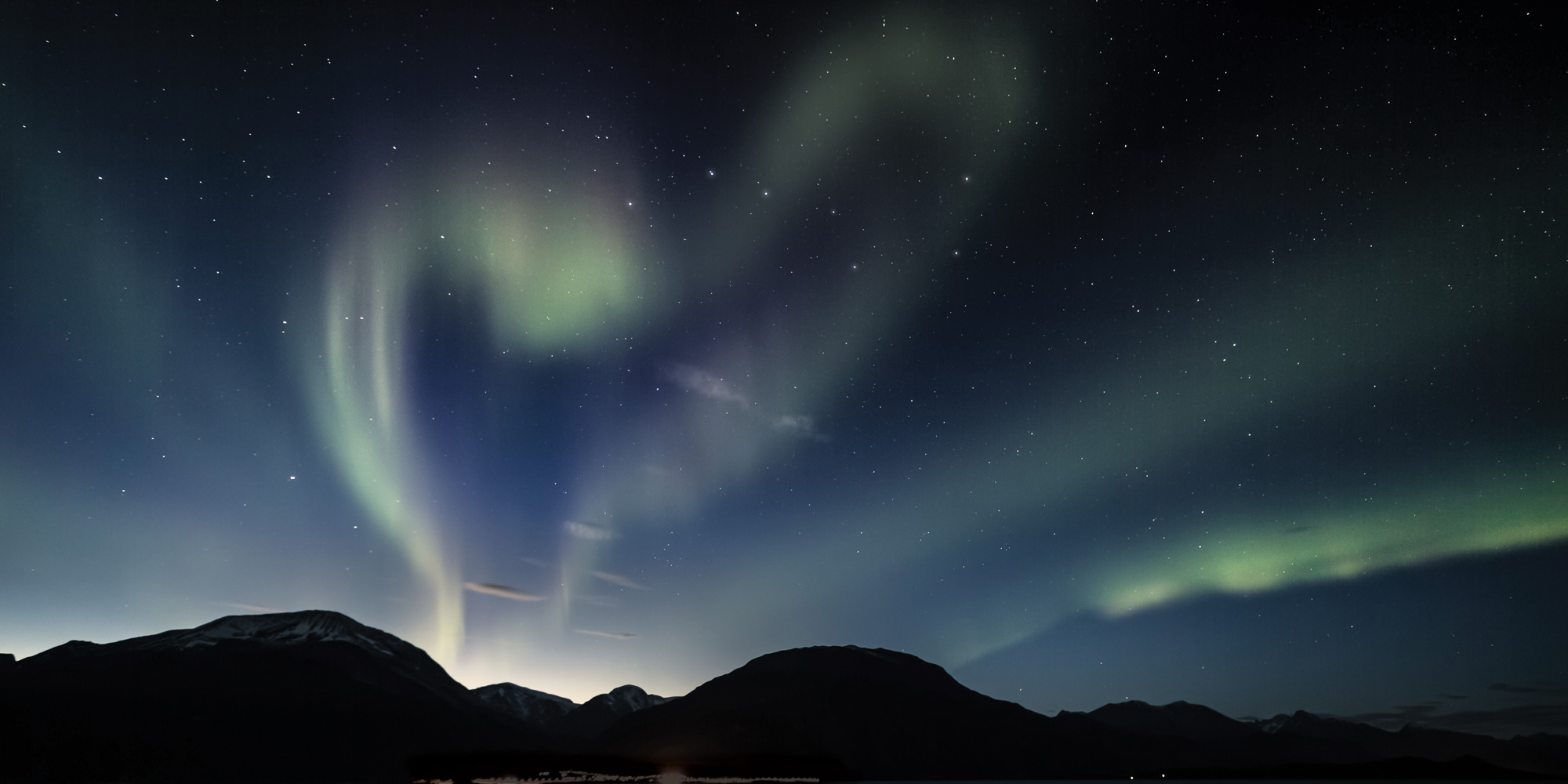 Green aurora borealis lights in a dark sky form the shape of a heart over a dark mountain landscape silhouette.