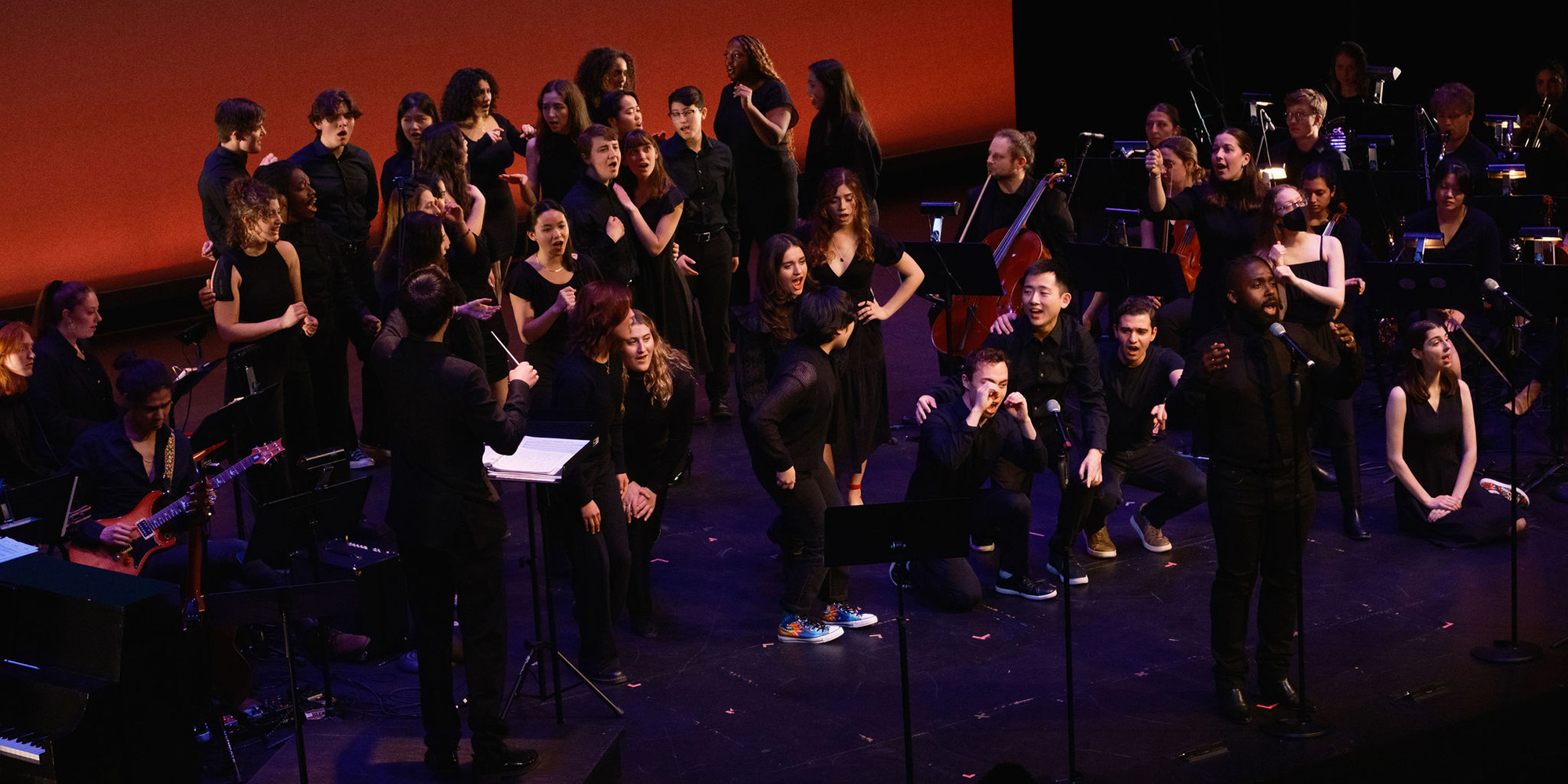 A large group chorus, all dressed in black, stands singing onstage amidst orchestra musicians on both sides.