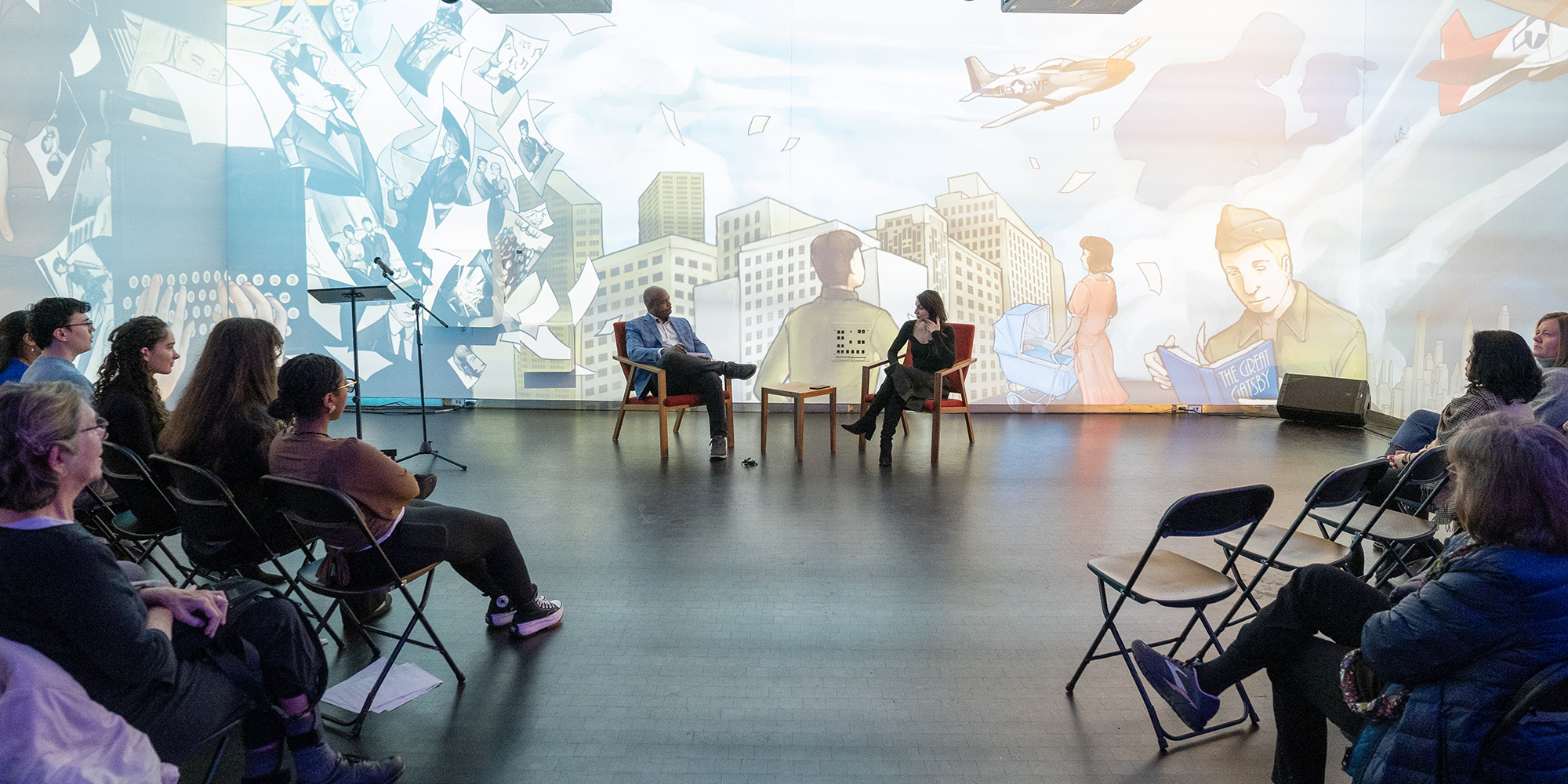 Two people sit in chairs having a discussion while a seated audience watches.