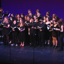 A choir stands onstage singing while musicians play, seated on the right, and a conductor leads on the left.