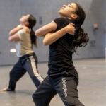 Two dancers hug their arms across their chests while standing with legs spread wide.