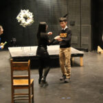 Two actors talk and interact in the middle of a theater space while 2 others are seen at their right and left.