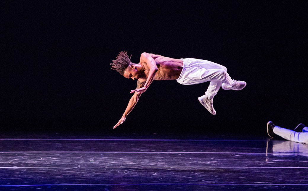 A dancer floats in the air after jumping, with their body horiztonal to the ground and arms out in front of them.