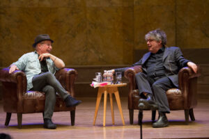 Two people sit in chairs talking on a stage.
