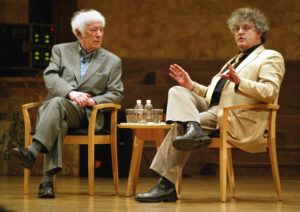 Paul Muldoon and Seamus Heaney sit in chairs to talk and gesture with their hands.