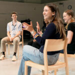 A group of students sit in chairs in a semi-circle in a studio.