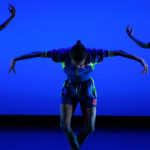 Three dancers with their arms held outright in various positions perform on stage.